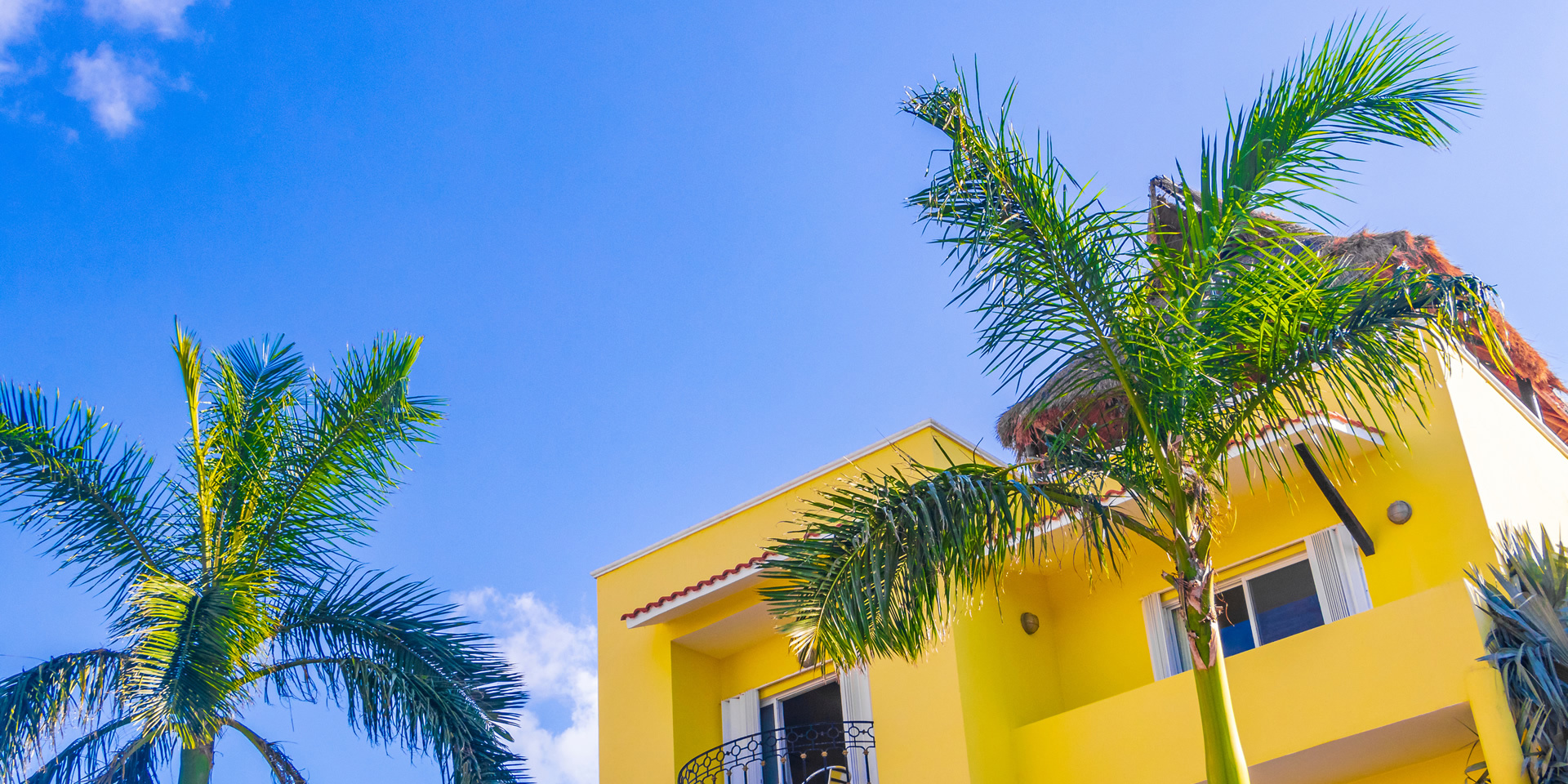 Yellow building with balconies and palm trees against a bright blue sky.