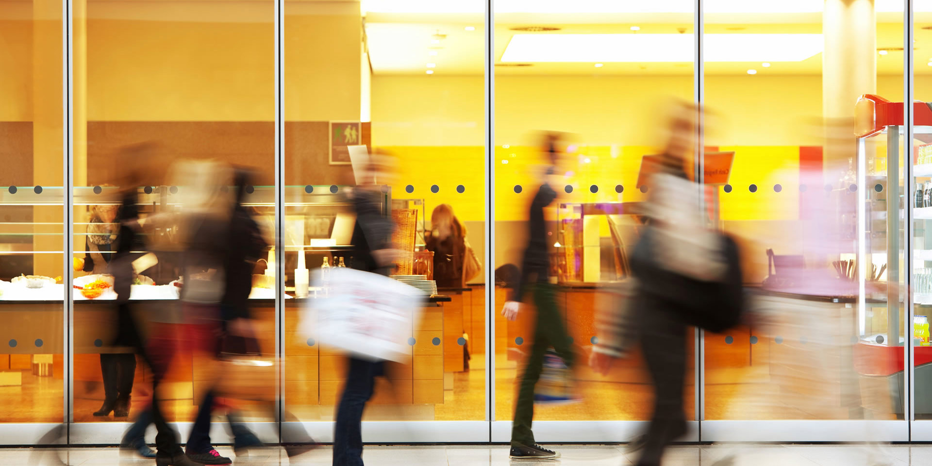 People walking past a glass‑walled café with warm yellow lighting, shown in motion blur inside a busy indoor space.