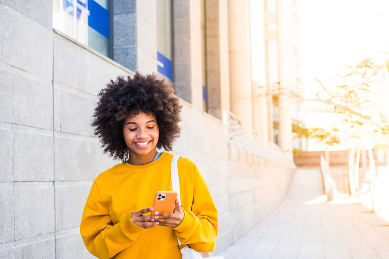 Person standing outdoors near a modern building, looking at a smartphone and carrying a shoulder bag.